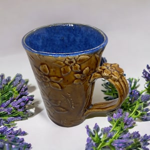 May include: A brown ceramic mug with a blue glaze inside and a floral design. The mug has a handle that is shaped like a flower.