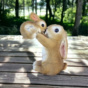 May include: A light brown bunny figurine with a smaller bunny held in its arms. The larger bunny is sitting down, with its arms raised to hold the smaller bunny. The bunnies have large, dark eyes and pink inner ears. The figurine is on a wooden surface.