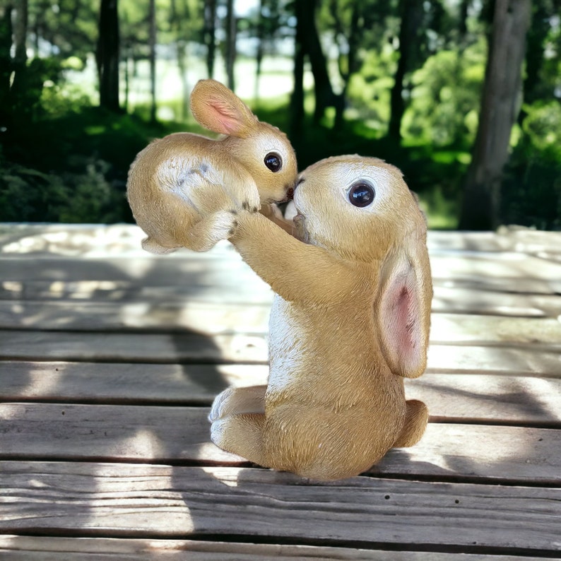 May include: A light brown bunny figurine with a smaller bunny held in its arms. The larger bunny is sitting down, with its arms raised to hold the smaller bunny. The bunnies have large, dark eyes and pink inner ears. The figurine is on a wooden surface.
