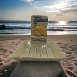 May include: A ceramic soap dish with a raised design of two hands washing under a faucet. The dish has a drain and the words "Wash Me" are printed on the front.