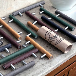 May include: An assortment of leather cabinet pulls in various colours, including brown, tan, green, grey, and black. The pulls have a stitched edge and are arranged on a light grey countertop. A brown paper tube with the text "Gaffe Leather" is visible in the centre of the image.