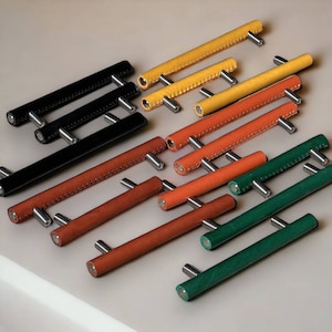 Könnte beinhalten: Sortiment an Leder-Schubladengriffen in verschiedenen Farben, darunter Schwarz, Braun, Gelb und Grün. Die Griffe haben ein genähtes Design und Metallakzente.
