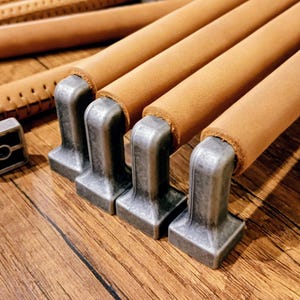 Op de afbeelding: Close-up van verschillende kastgrepen met een lichtbruin lederen handvat en een vierkante, antiek ogende metalen basis. De grepen zijn gerangschikt op een houten oppervlak, wat hun ontwerp en textuur laat zien.