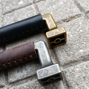 May include: Two leather wrapped handles with metal ends. The ends are square shaped with a round hole in the center. One handle is brown leather with a silver end and the other is black leather with a gold end.