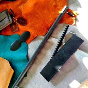 Peut inclure: Un ensemble d'outils et d'accessoires de maroquinerie. L'image présente une longue règle en cuir noir avec une poignée en métal, un étui en cuir noir et un outil en cuir noir. Diverses pièces de cuir dans les tons marron et bleu sont en arrière-plan.