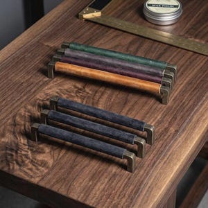 Puede incluir: Una colección de seis tiradores de armario con agarres cilíndricos envueltos en cuero. Tres tiradores tienen cuero de colores verde, morado y tostado, mientras que tres tienen texturas de ante gris oscuro. Todos tienen soportes metálicos en L.