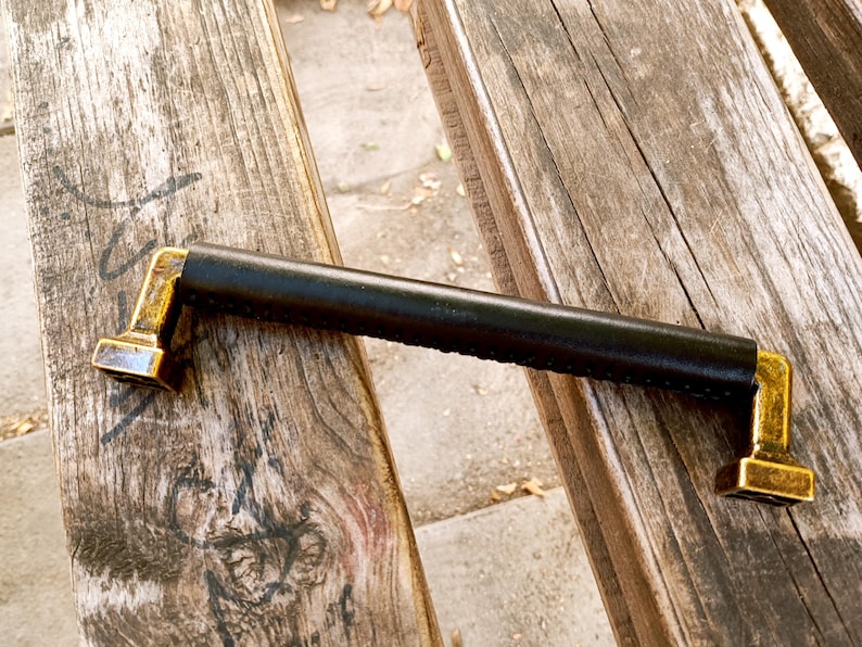 May include: A black leather handle with gold metal ends. The handle is on a wooden surface.
