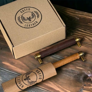 May include: Two cabinet pulls with leather-wrapped handles in dark brown and tan, featuring brass-toned metal ends. One pull is partially encased in a branded cardboard sleeve. A matching cardboard box displays the GAFFO LEATHER logo.