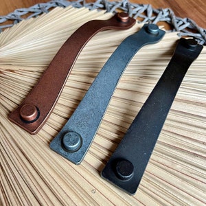 Puede incluir: Tres tiradores de armario de cuero en diferentes acabados: cobre, gris azulado y negro. Cada tirador tiene una forma rectangular redondeada con un pomo redondo en el extremo.