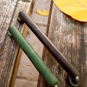 May include: Two cylindrical cabinet pulls. One is dark brown, the other is green. Both have a metal base and are resting on a weathered wooden surface. A yellow leaf is in the background.