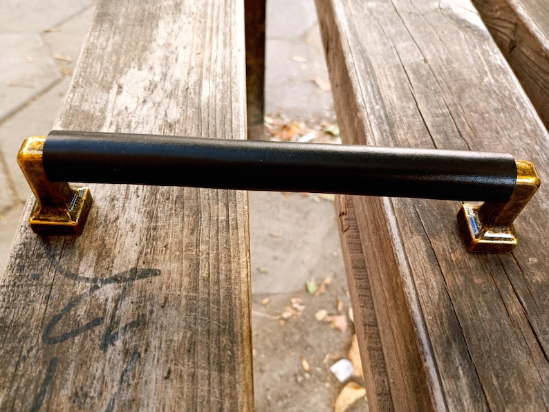 May include: Black metal door handle with gold accents. The handle is rectangular and has a smooth, flat surface.