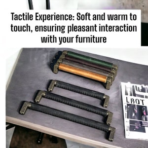Peut inclure: Cinq poignées de tiroir en cuir avec des ferrures en laiton. Les poignées sont de différentes nuances de brun, y compris le brun foncé, le beige et le brun clair. Les poignées sont disposées en rangée sur une surface grise. Le texte "Tactile Experience: Soft and warm to touch, ensuring pleasant interaction with your furniture" est au-dessus des poignées.