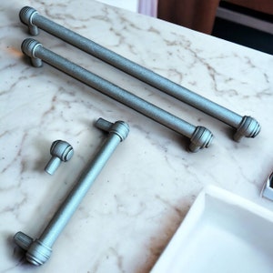 Può includere: Tre tiretti per cassetti in metallo argentato con un design arrotondato. I tiretti sono su una superficie di marmo bianco.