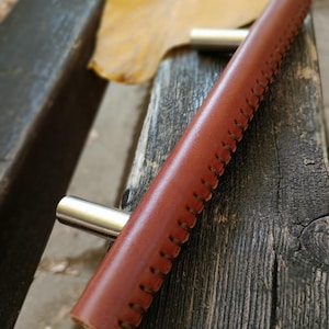 Op de afbeelding: Een bruine leren kastgreep met een gestikte rand en zilveren metalen hardware.