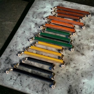 May include: A set of 10 leather cabinet pulls in various colors, including black, yellow, green, brown, and orange. The pulls are all the same size and shape, with a simple, modern design.