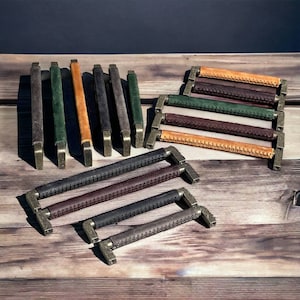 May include: Assortment of leather drawer pulls in various colors, including brown, green, and black. The pulls have a stitched design and are mounted on metal brackets.