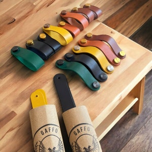 Könnte beinhalten: Sortiment an Leder-Schubladengriffen in verschiedenen Farben, darunter Schwarz, Grün, Gelb und Braun. Die Griffe werden auf einer Holzoberfläche mit dem Markennamen "GAFFO" auf zwei Pappröhren präsentiert.