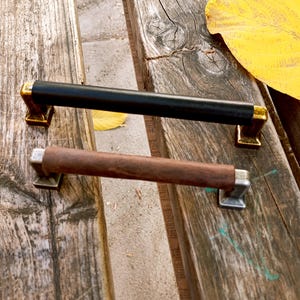 May include: Two cabinet door pulls, one black and one brown wood. The black pull has gold accents. The brown pull has silver accents.