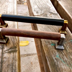 Puede incluir: Dos tiradores para puertas de armario, uno con un tirador de cuero negro y detalles dorados, y el otro con un tirador de madera marrón y detalles plateados.