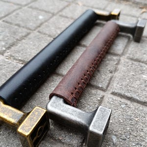 Op de afbeelding: Twee metalen kastgrepen met leren banden. De grepen zijn messingkleurig en hebben een vierkante basis. De leren banden zijn bruin en zwart.