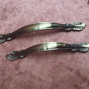 May include: A pair of antique bronze-effect cabinet handles. They feature a curved design with decorative, scrolled ends. The handles are set against a pinkish-brown background. These decorative pulls are likely for furniture.