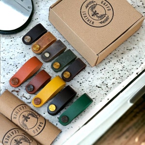 Op de afbeelding: Assortiment van tien leren snoerorganizers in verschillende kleuren, waaronder bruin, groen, geel en zwart. Elke organizer heeft een messing metalen lus en is verpakt in een bruine doos met de tekst "Gaffeo Leather" erop gedrukt.