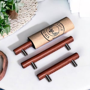 Op de afbeelding: Drie bruine lederen kastgrepen met zilveren beslag. De grepen zijn verpakt in een bruine papieren buis met de tekst "GAFFO LEATHER" erop gedrukt.