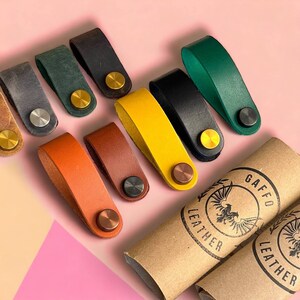 Könnte beinhalten: Sortiment an bunten Leder-Kabelbindern mit Messing- und Silberakzenten. Die Kabelbinder sind auf einem rosa Hintergrund mit zwei braunen Pappröhren mit der Aufschrift "GAFFO LEATHER" angeordnet.