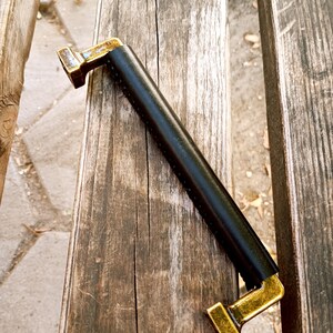 May include: Black leather cabinet pull with gold metal end caps. The pull is on a wooden surface.