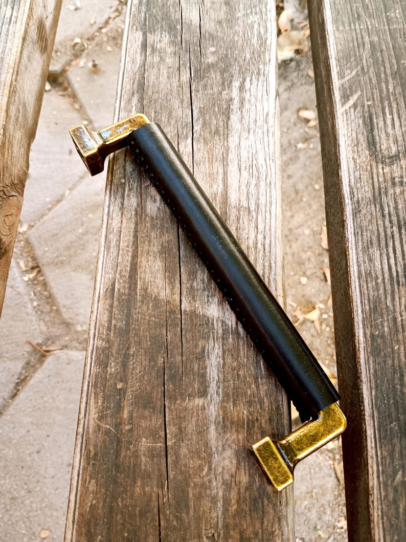 May include: Black leather cabinet pull with gold metal end caps. The pull is on a wooden surface.