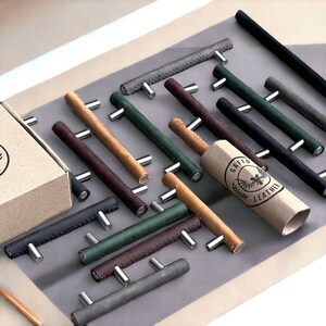 May include: An assortment of leather drawer pulls in various colours, including brown, black, green, and grey. The pulls have a stitched design and are arranged on a grey surface. A brown paper tube with the text "Gaffeo Leather" is visible in the image.