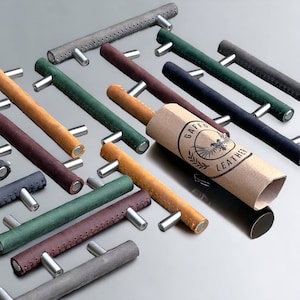 Op de afbeelding: Een assortiment lederen kastgrepen in verschillende kleuren, waaronder bruin, groen, grijs en bordeauxrood. De grepen hebben een gestikte rand en zijn gerangschikt op een grijs oppervlak. Een bruine kartonnen buis met de tekst "Gaffe Leather" bevindt zich in het midden van de afbeelding.