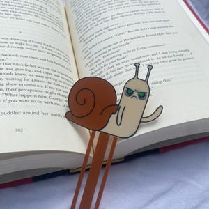 May include: A brown and beige snail bookmark with a grumpy expression. The snail is holding a brown bookmark with a metal clip. The bookmark is on top of an open book with text.