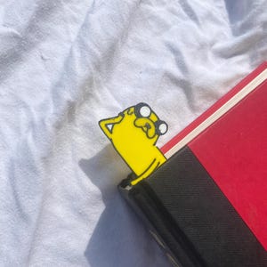 Peut inclure: Signet de livre en forme de chien jaune de dessin anim&eacute; avec des yeux noirs et un contour noir. Le signet sort d'un livre rouge et noir.