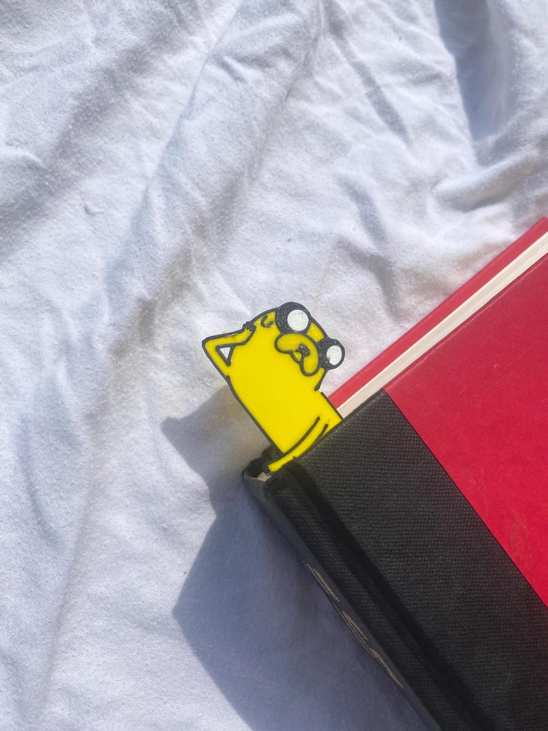 Peut inclure: Signet de livre en forme de chien jaune de dessin anim&eacute; avec des yeux noirs et un contour noir. Le signet sort d'un livre rouge et noir.