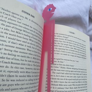 May include: Pink plastic bookmark in the shape of a stylized figure with a single blue eye, placed in an open book. The bookmark is a solid pink color and has a simple, cartoonish design.