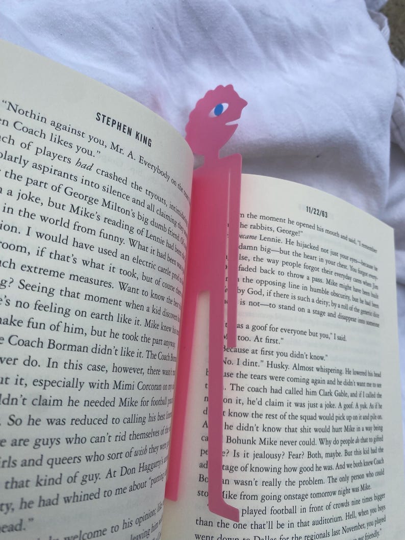 May include: Pink plastic bookmark in the shape of a stylized figure with a single blue eye, placed in an open book. The bookmark is a solid pink color and has a simple, cartoonish design.