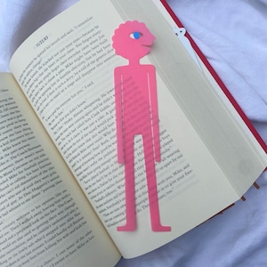May include: Pink die-cut bookmark in the shape of a person with a blue eye, placed in an open book. The bookmark has a scalloped head and a simple, elongated body with feet. The book's pages are visible, with text and red edges.