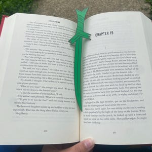 May include: A green paper bookmark in the shape of a sword is inserted into a book. The bookmark is made of folded paper and has a pointed tip. The book is open to Chapter 19.