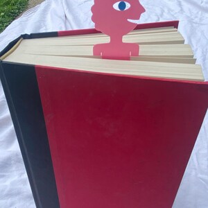 May include: A large red book with a black spine and a pink bookmark. The bookmark is a stylized human figure with a blue eye. The book's pages are cream-colored, and the cover is a glossy red.