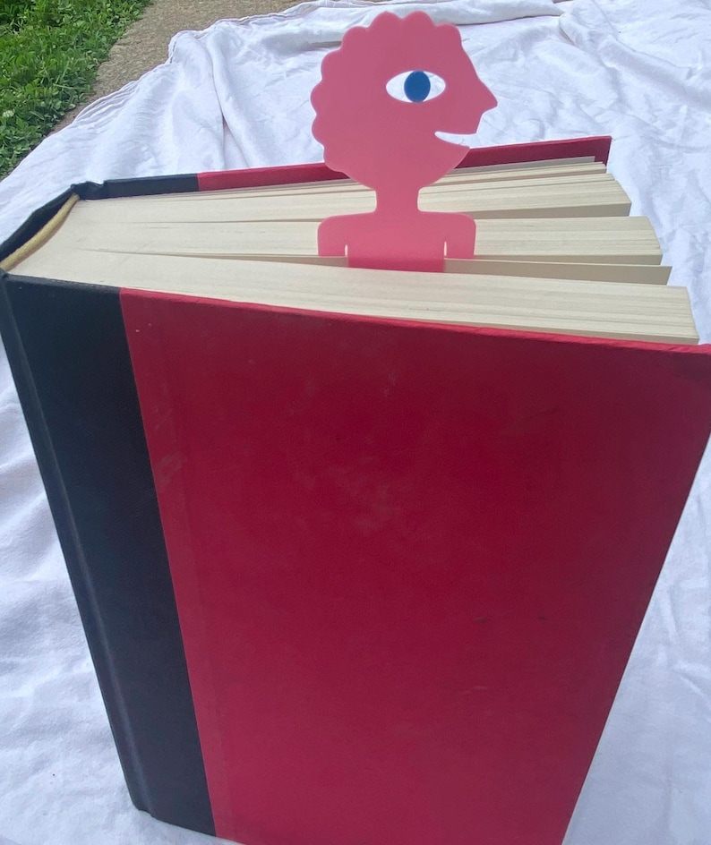 May include: A large red book with a black spine and a pink bookmark. The bookmark is a stylized human figure with a blue eye. The book's pages are cream-colored, and the cover is a glossy red.