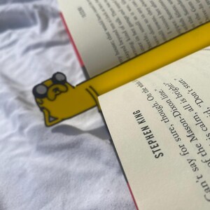 Peut inclure: Un marque-page jaune et noir repr&eacute;sentant un chien de dessin anim&eacute; avec un nez rouge et des lunettes. Le marque-page est plac&eacute; dans un livre avec le texte "Stephen King" sur la couverture.