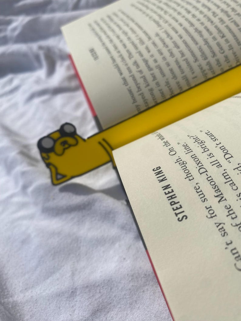 Peut inclure: Un marque-page jaune et noir repr&eacute;sentant un chien de dessin anim&eacute; avec un nez rouge et des lunettes. Le marque-page est plac&eacute; dans un livre avec le texte "Stephen King" sur la couverture.