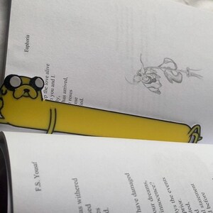 Peut inclure: Un marque-page jaune en forme de chien de dessin anim&eacute; avec des yeux noirs et un nez noir. Le marque-page est plac&eacute; sur un livre ouvert avec du texte sur les pages. Le texte sur la page est "F.S. Yousaf" et "Euphoria".