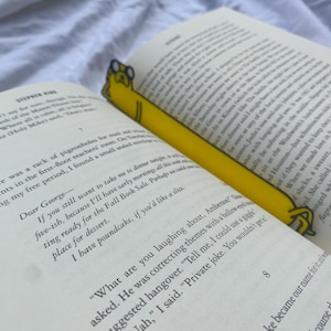 Peut inclure: Un marque-page jaune en forme de chien de dessin anim&eacute; avec un livre dans sa gueule. Le marque-page est plac&eacute; dans un livre ouvert avec du texte sur les pages. Le texte dit "Cher George - Si tu veux toujours m'emmener d&icirc;ner ce soir vers cinq heures, parce que j'aurai des matins..."
