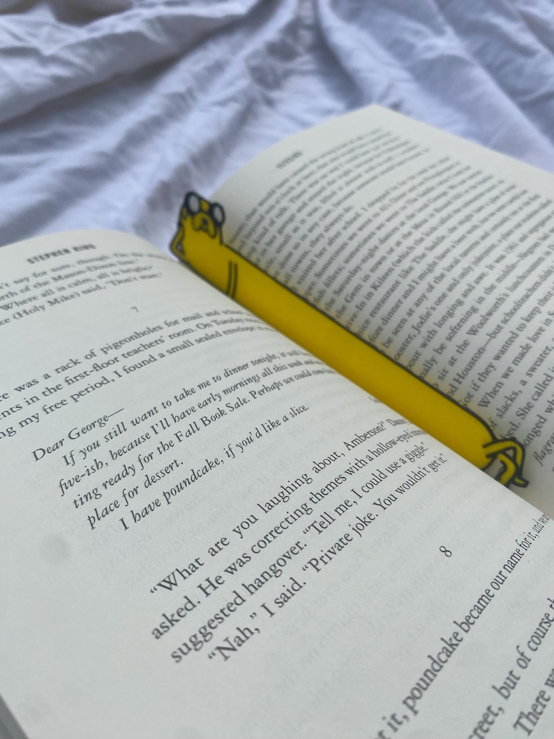 Peut inclure: Un marque-page jaune en forme de chien de dessin anim&eacute; avec un livre dans sa gueule. Le marque-page est plac&eacute; dans un livre ouvert avec du texte sur les pages. Le texte dit "Cher George - Si tu veux toujours m'emmener d&icirc;ner ce soir vers cinq heures, parce que j'aurai des matins..."