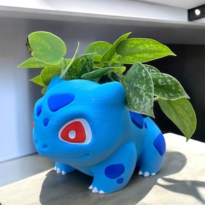 May include: A blue Bulbasaur plant pot with a red and white eye design, holding green leafy plants. The pot has blue spots and white feet. The pot is made of a hard material and is sitting on a light-coloured surface.