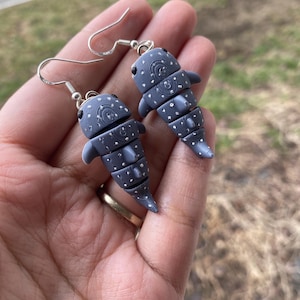 May include: A pair of silver dangle earrings featuring a gray and white whale shark design. The earrings are made of polymer clay and have a detailed, textured finish.