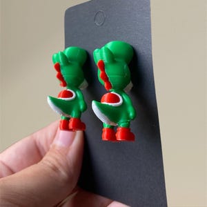 Pode incluir: Um par de brincos Yoshi verdes e vermelhos. Os brincos estão presos a um cartão de exibição preto. Os brincos são um design impresso em 3D do personagem Yoshi da série de videogames Super Mario.