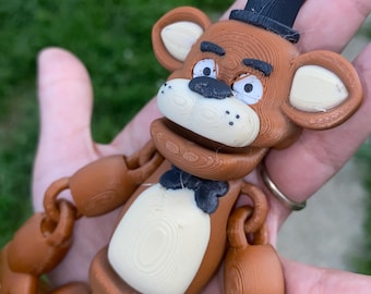 May include: A brown articulated Freddy Fazbear figure with a black top hat and bow tie. The bear has white ears, muzzle, and teeth. The figure is made of plastic and has visible layer lines from the 3D printing process.